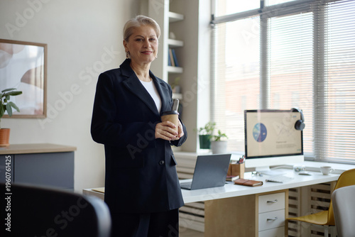 Portrait of senior businesswoman standing in office, holding takeaway coffee and looking at camera. Suitable for leadership, executive branding, mentoring, career growth marketing