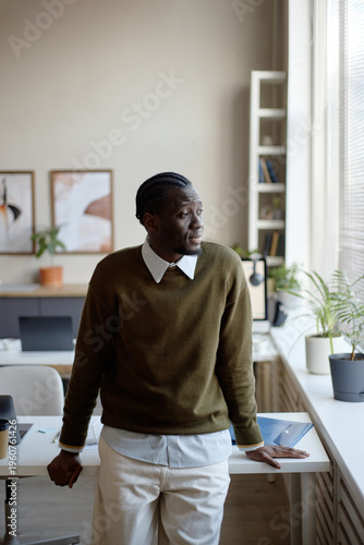 Vertical shot of Black young adult man leaning on desk and looking through office window, reflecting on work strategy. Suitable for business planning, leadership, career growth messaging