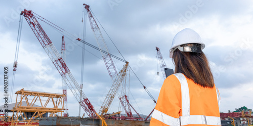 A worker stands on a construction site. Several cranes are in operation, lifting materials. The sky is cloudy, and the worker looks at the ongoing tasks with a focus on safety