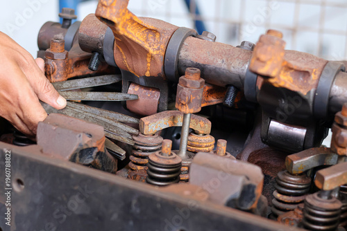 фотографія ​A mechanic's hand uses a feeler gauge to measure valve clearance on a rusty heavy-duty diesel engine cylinder head inside a workshop during a maintenance process