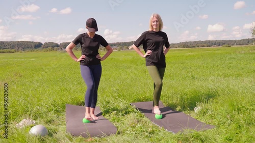 Two active women performing foot massage and proprioception exercises with hemisphere balance trainers on yoga mats in a sunlit green field, improving coordination and strengthening ankle muscles