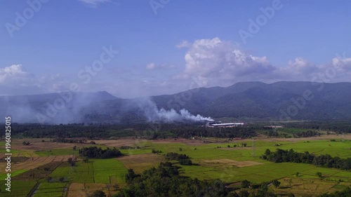 Air pollition releasing from factory buildings at the feet of large mountain formation, with lush tropical foliage and sunny clear sky in the background.