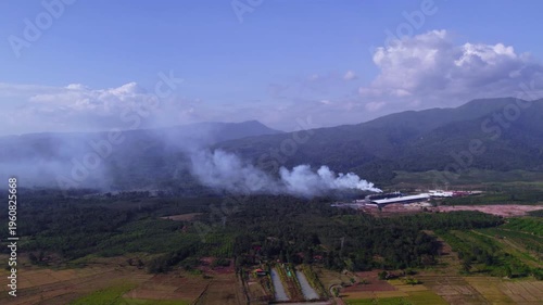 Aerial view of air pollition releasing from factory buildings at the feet of large mountain formation, with lush tropical foliage and sunny clear sky in the background.