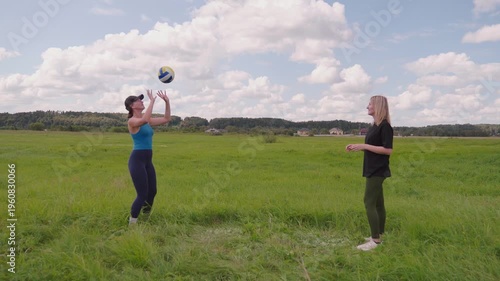 Two athletic women enjoying a beautiful summer day playing volleyball in a vast green field with a blue sky and white clouds, throwing and catching the ball in a friendly match