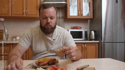 White chubby obese man with beard sitting at kitchen table eating homemade dish of meat, potatoes and fresh vegetables while enjoying lunch or dinner with appetite
