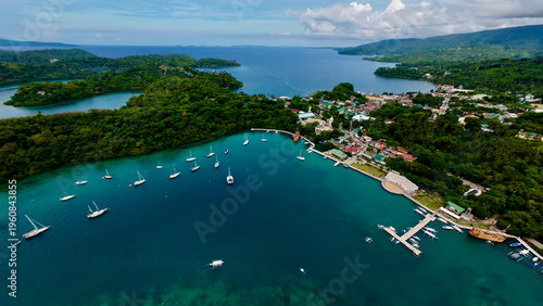 Aerial view of Puerto Galera bay with turquoise water and anchored boats. Lush green hills surround the picturesque coastal town under bright sky.