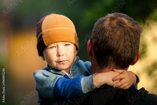 Little boy in a hat being held by an adult, looking at the camera with soft light and a blurred outdoor background.