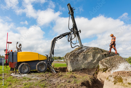 Male worker standing near machine digging large rock with hydraulic top hammer drill against cloudy sky