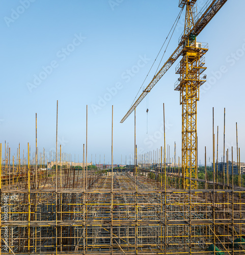 Yellow tower crane working on a large-scale building construction site with extensive scaffolding under clear blue sky.