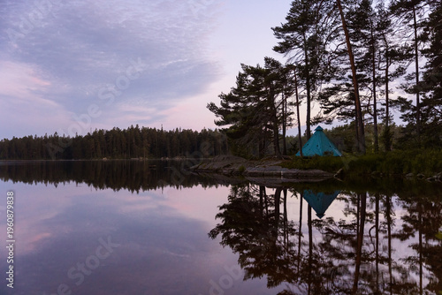 Canvas Print A tranquil, early morning photograph of a teal tipi tent pitched amongst pine trees on a rocky shore, its perfect reflection on the mirror-like surface of a calm Swedish lake at dawn