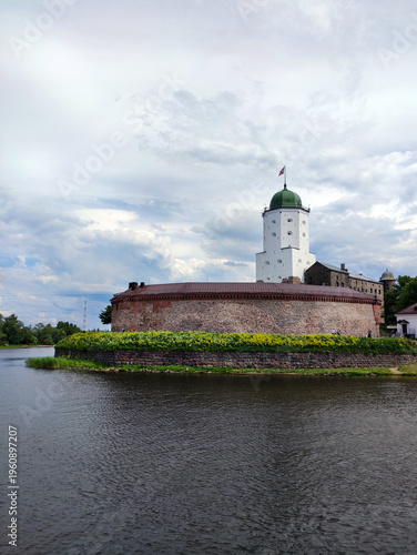 Overview of the castle on an island at the Vyborg Combined Museum-Reserve, featuring a white tower with green dome and flag, stone walls, calm water. June 6, 2024.
