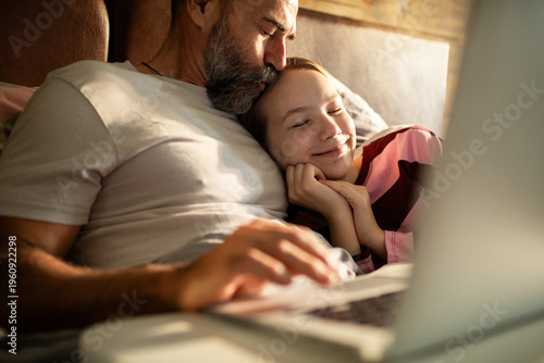 Father kissing daughter while cuddling in bed with laptop at home