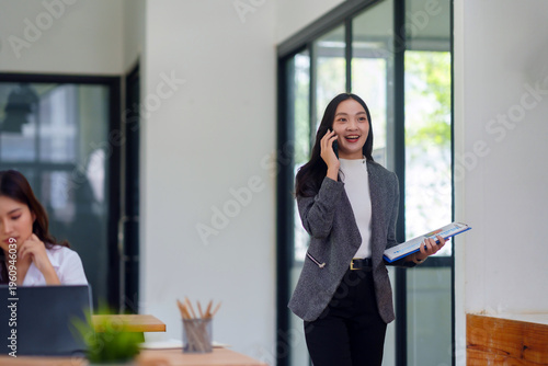 Smiling Asian businesswoman walking confidently, talking on phone with document, showing career success and professional communication