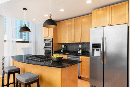 A contemporary kitchen features light wood cabinets, black granite countertops, a black subway tile backsplash, and a large island with bar stools. No brands or labels.