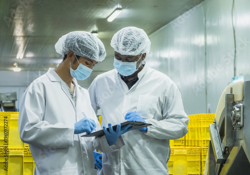 Foto Two diverse quality control inspectors in protective gear discussing production data on a clipboard in a food manufacturing plant to maintain global compliance and operational excellence