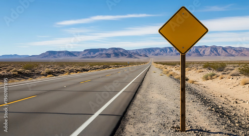 Empty desert highway with yellow warning sign under a vast blue sky with distant mountains