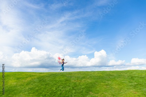 Under the blue sky and white clouds, a little girl is playing with a balloon on the grass