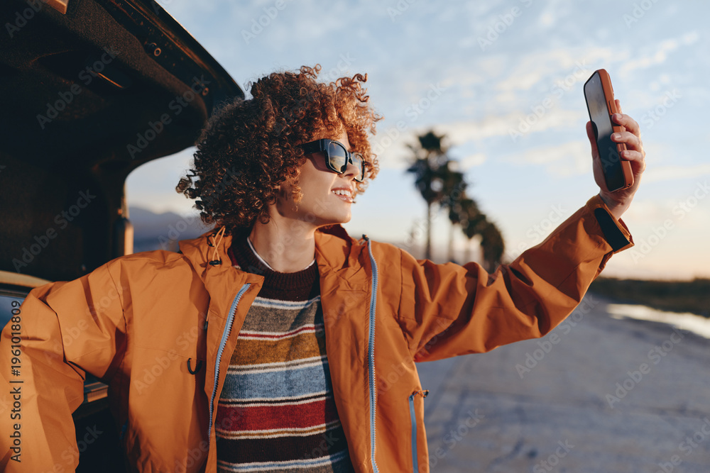 Obraz premium Smiling woman with curly hair wearing rainbow sweater and orange jacket taking selfie with smartphone near car outdoors. Lifestyle concept with casual clothing and sunny weather.