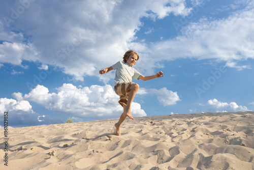 preteen boy runs energetically barefoot along a sandy beach on a sunny summer day under a blue sky with white clouds. A happy, carefree childhood, long-awaited holidays