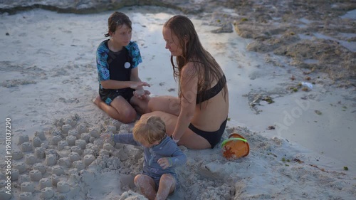 Young mom spending quality time with her two sons on a beautiful tropical beach, enjoying sunshine, laughter, and family bonding moments by the sea.