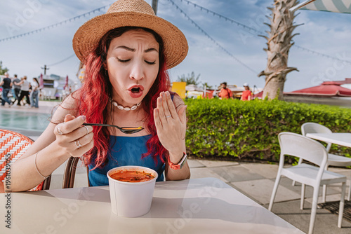Young woman with red hair and straw hat showing a funny facial expression after tasting hot spicy street food soup