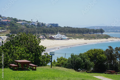 South Africa,Mossel Bay,Bartolomeu Dias Museum Complex,A scenic view of a sandy beach with clear blue water, surrounded by greenery and a picnic area in the foreground.