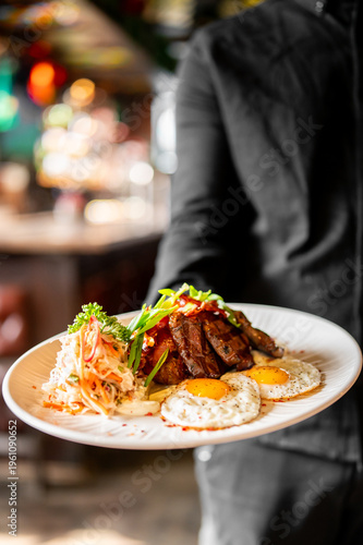 Foto A waiter's hand holds a plate of grilled steak, two fried eggs, and fresh colesl