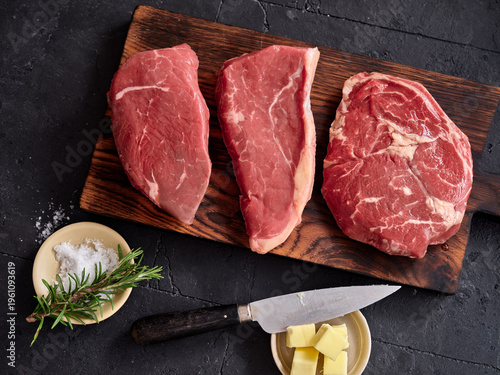 Raw rib-eye, roast beef, flank and rump steak on a wooden board with knife and butter at a kitchen counter