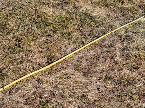 Yellow garden hose lying diagonally on dry grass with fallen leaves in early spring or autumn. Outdoor natural background with seasonal texture