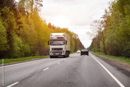 Large white semi truck driving on asphalt highway road through green forest area under bright sun light during daytime travel
