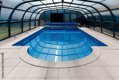 Slika na platnu Wide view of an indoor swimming pool beneath a transparent enclosure with tiled deck and calm blue water