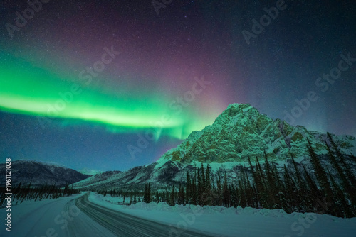 View of the Aurora Borealis dances above the snow-covered road and majestic, snow-capped mountain peaks under a starlit sky, Coldfoot, Alaska, United States.
