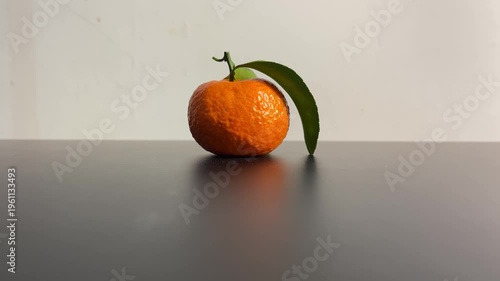 Fresh ripe tangerine with green leaf on a dark surface in a minimal setting. Natural light highlights the texture of the citrus fruit, creating a clean and calming still life composition.
