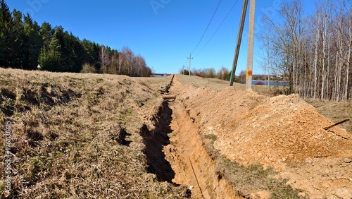 Fotografía A trench has been dug along the edge of the meadow along an asphalt road and a power line for laying pipelines and cables