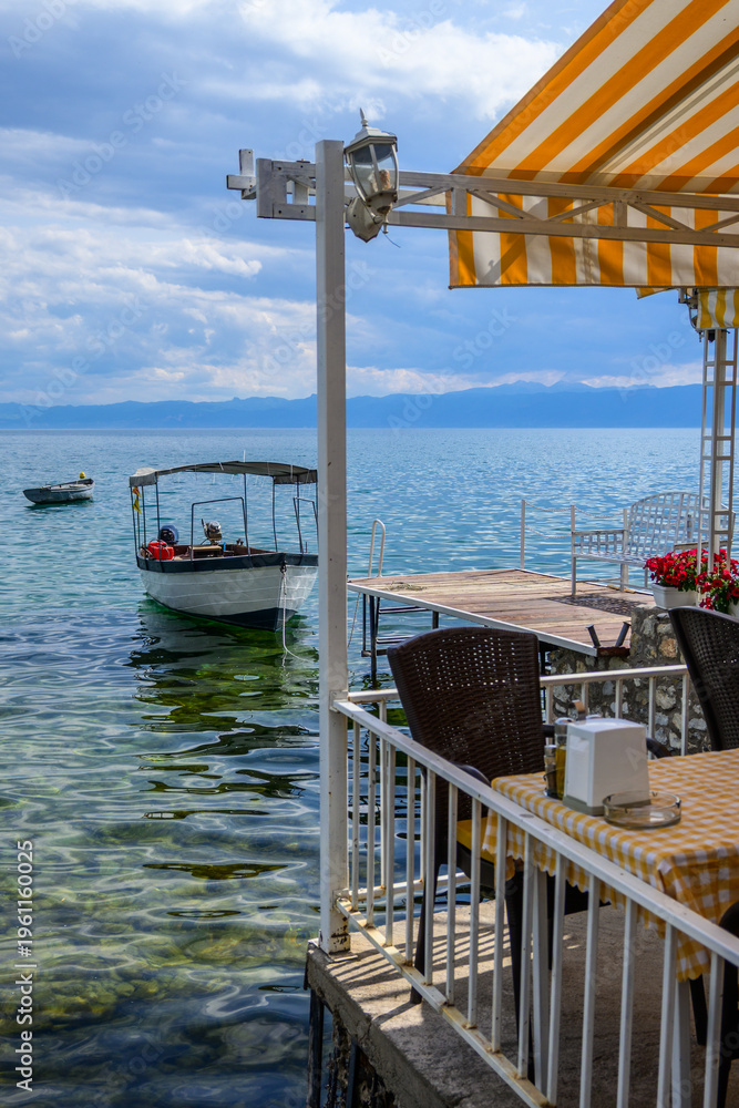 Fototapeta premium A sunny restaurant terrace with striped awning and outdoor seating offers views of Lake Ohrid, where boats gently float on the blue-green water.