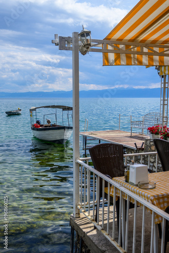A sunny restaurant terrace with striped awning and outdoor seating offers views of Lake Ohrid, where boats gently float on the blue-green water.