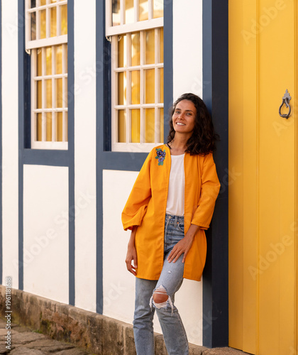 Woman stands near yellow door wearing orange jacket in a colorful neighborhood during daylight hours