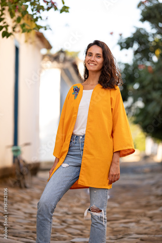 Woman wears orange outerwear while posing on a cobblestone street with trees and buildings in the background during daytime