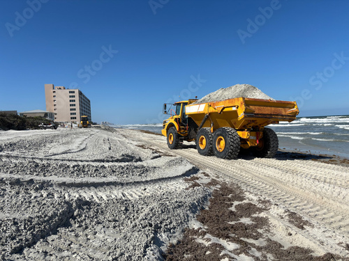 Dump truck placing sand on Daytona Beach, Florida during coastal protection and beach renourishment project after storm erosion, with ocean waves and clear blue sky in background.