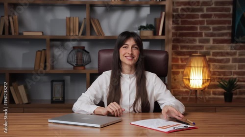 Business woman closes laptop and smiles while sitting at a desk in an office setting.