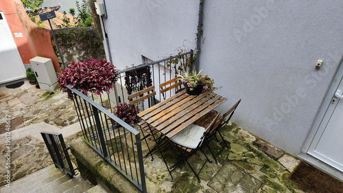 Small outdoor terrace with wooden table, folding chairs and potted plant near white wall. Cozy urban patio scene suitable for lifestyle content, home decoration ideas and small space design concepts.