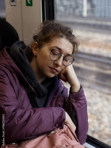 Young woman sleeping by train window during travel. Passenger resting with head on hand in carriage seat. Concept of fatigue, commute, journey, railway transport, relaxation and everyday lifestyle.