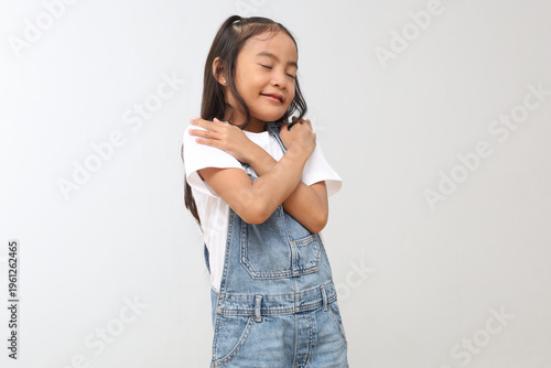 Happy and smiling Asian little girl hugging herself while closing her eyes, love yourself, be confident, feel proud, peaceful and relieved, mental health and self-respect concept