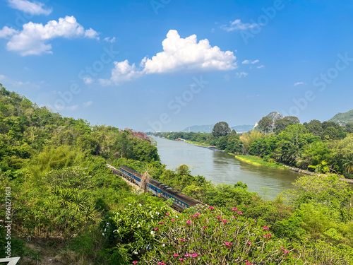 Train Running Along River with Mountain Landscape and Blue Sky in Thailand