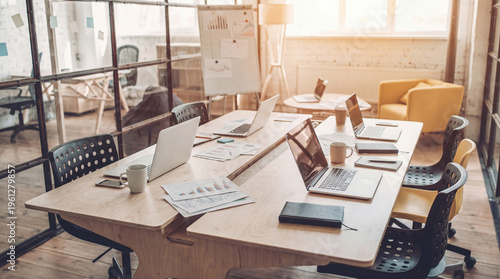 Photos Vibrant collaborative workspace with multiple laptops and documents on a large wooden table in a sunlit office