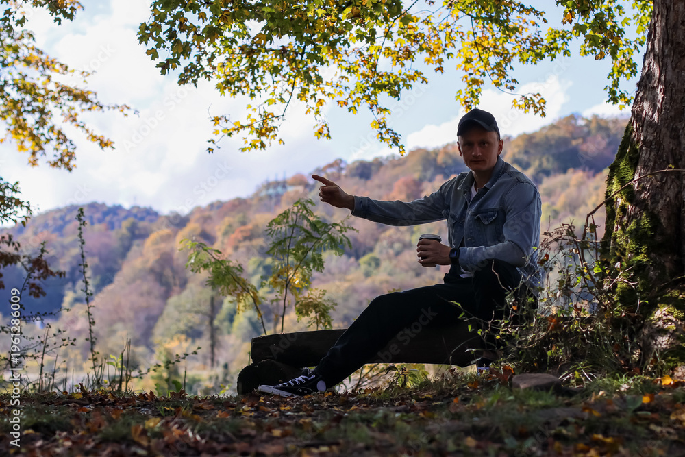 Fototapeta premium Male relaxing with beverage in fall forest landscape. Man in light blue jacket sits on wooden log sipping from cup amid yellow autumn leaves and mountainous backdrop.