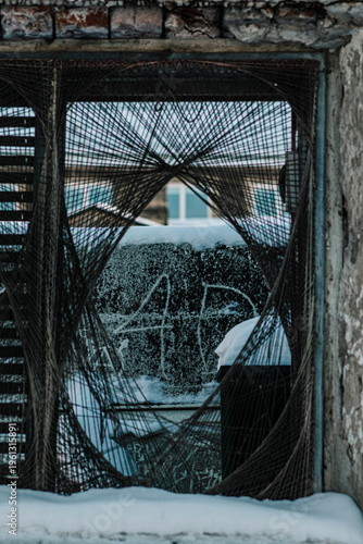 The image captures a view through a rugged window frame intricately woven with strings or wires, creating a unique, spiderweb-like pattern. In the background, a snowy urban setting with graffiti-cover