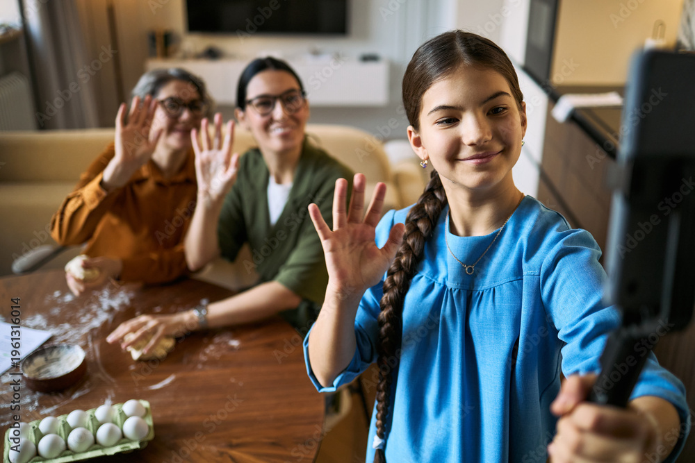 Obraz premium Teenage girl filming family baking video on smartphone, waving with mother and senior grandmother while preparing dough. Useful for family vlog, social media, parenting content marketing