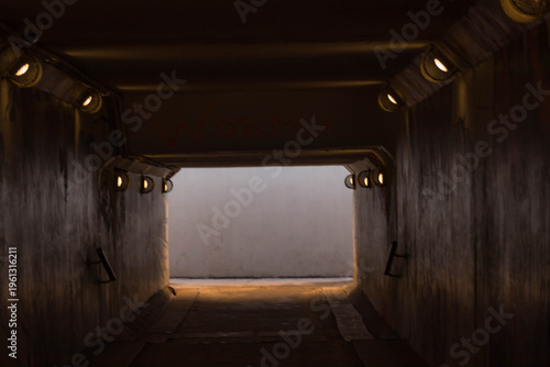The image captures a perspective from inside a dark pedestrian tunnel, its sides lined with rhythmically placed lights. The warm glow contrasts with the rough walls and the bright, clear exit at the f