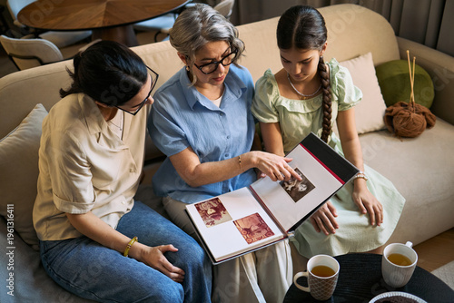 фотографія Senior woman with adult daughter and girl reviewing family photo album, sharing memories at home